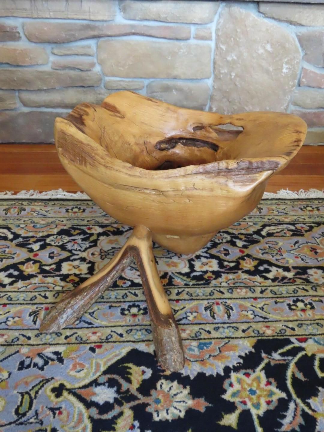 Wooden bowl on tripod base, on patterned rug, against a stone wall.