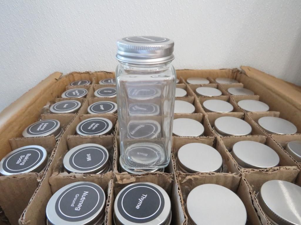 A glass spice jar standing in a cardboard box of empty spice jars with metal lids.