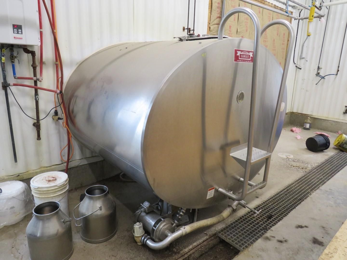 Large stainless steel milk tank with ladder and pump, flanked by metal milk cans in a dairy setting.