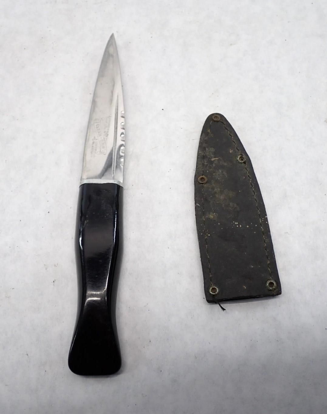 A silver knife with a black handle and a black sheath on a white surface.