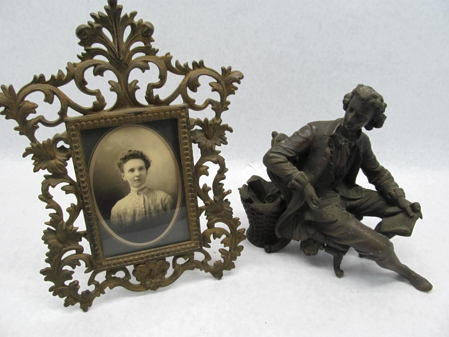 Ornate metal photo frame with oval portrait next to a bronze-colored statue of a seated man.
