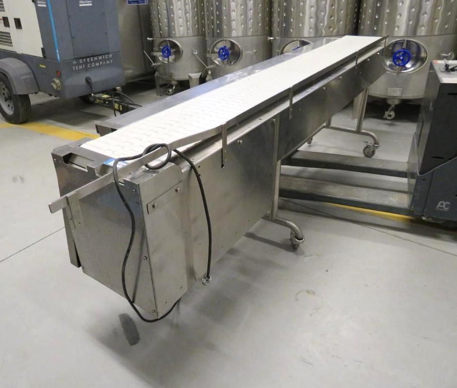 Stainless steel conveyor belt in a processing facility, with large tanks in the background.