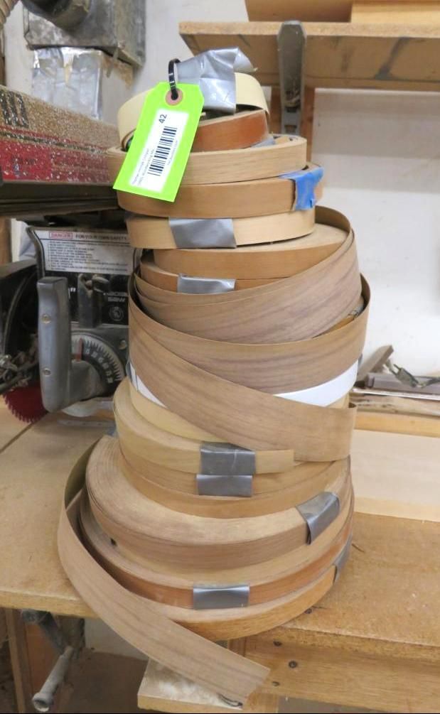 Spools of wood edge banding stacked on a workbench, various colors and widths, with metal clips and a tag.