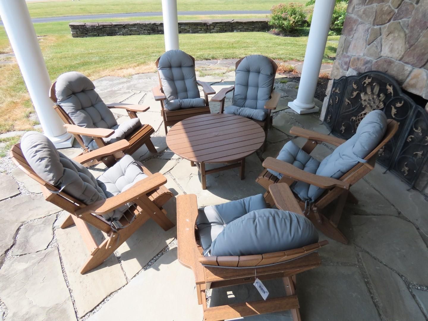 Patio seating arrangement: six wooden chairs with gray cushions around a round table on a stone patio.