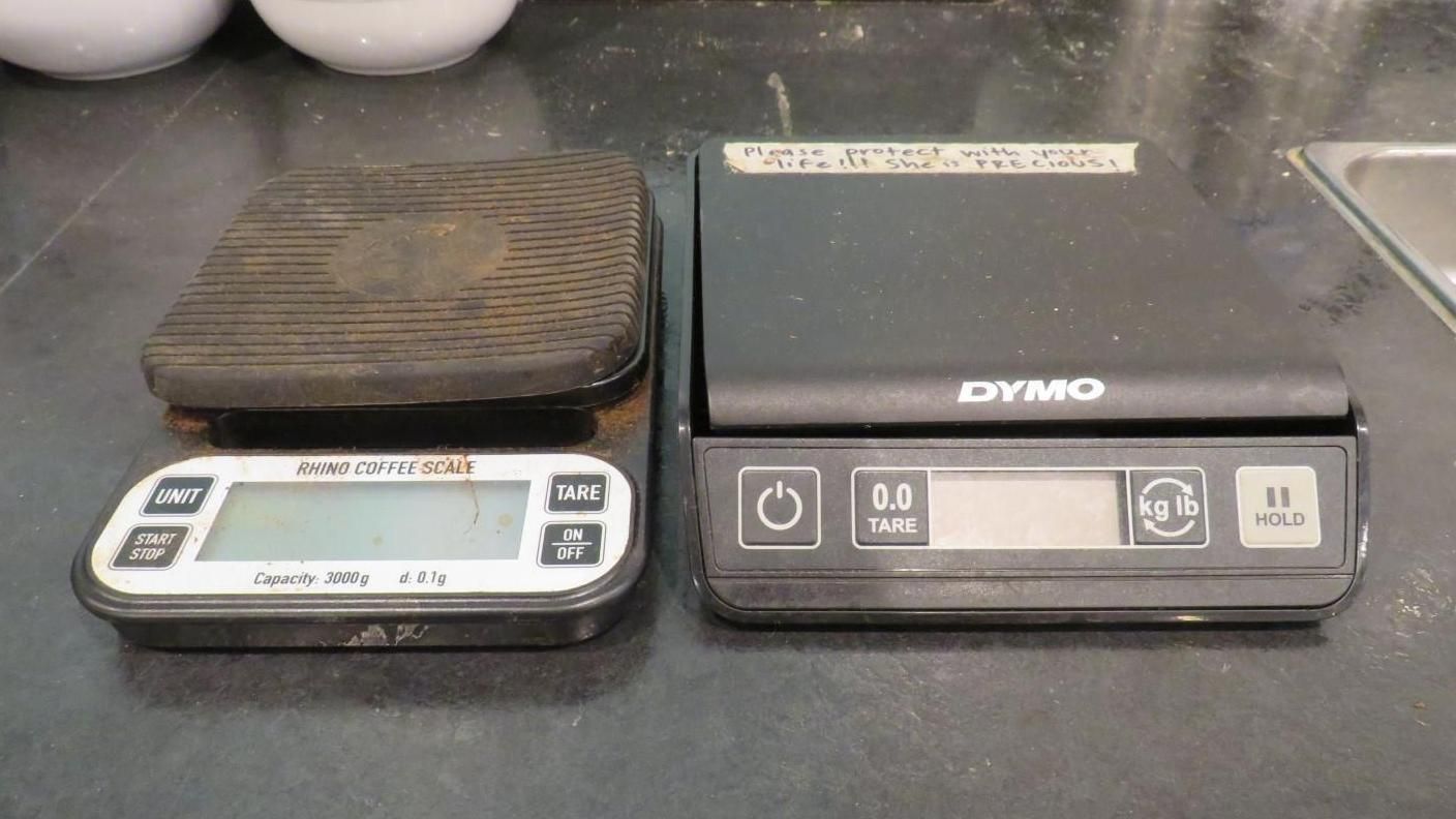 Two digital scales: one small, wooden-topped scale next to a larger, black Dymo scale, on a dark countertop.