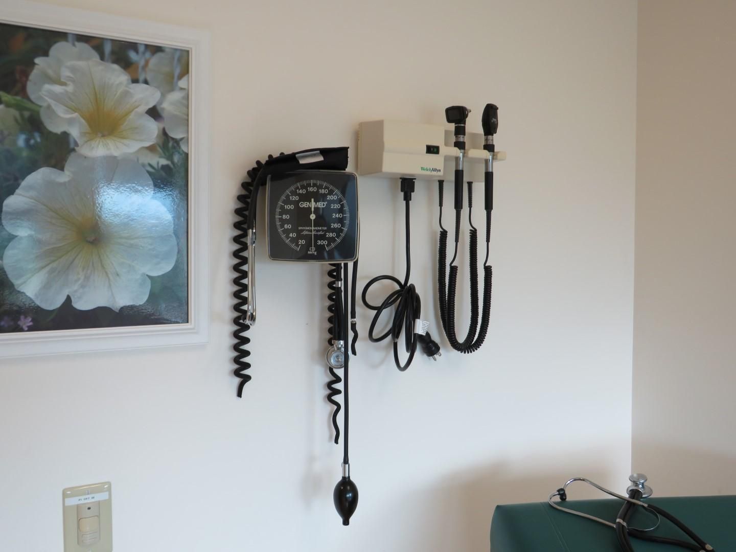 Blood pressure monitor and otoscope mounted on a wall in a medical office next to a framed floral picture.
