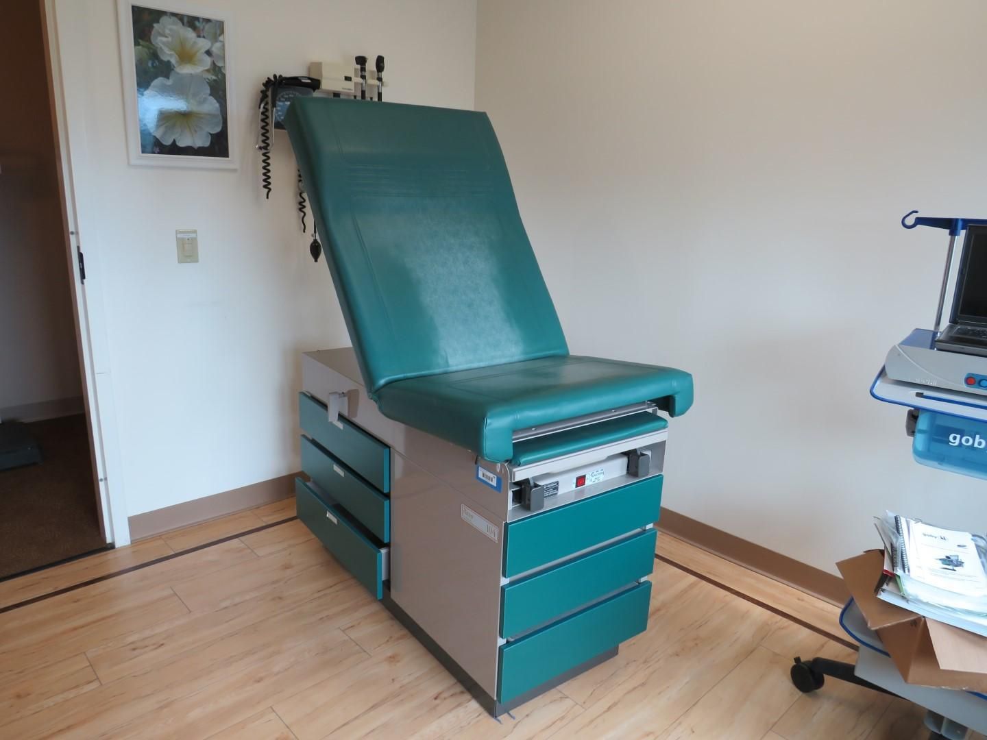 Examination room with green medical chair and drawers, white walls, and wood floor.