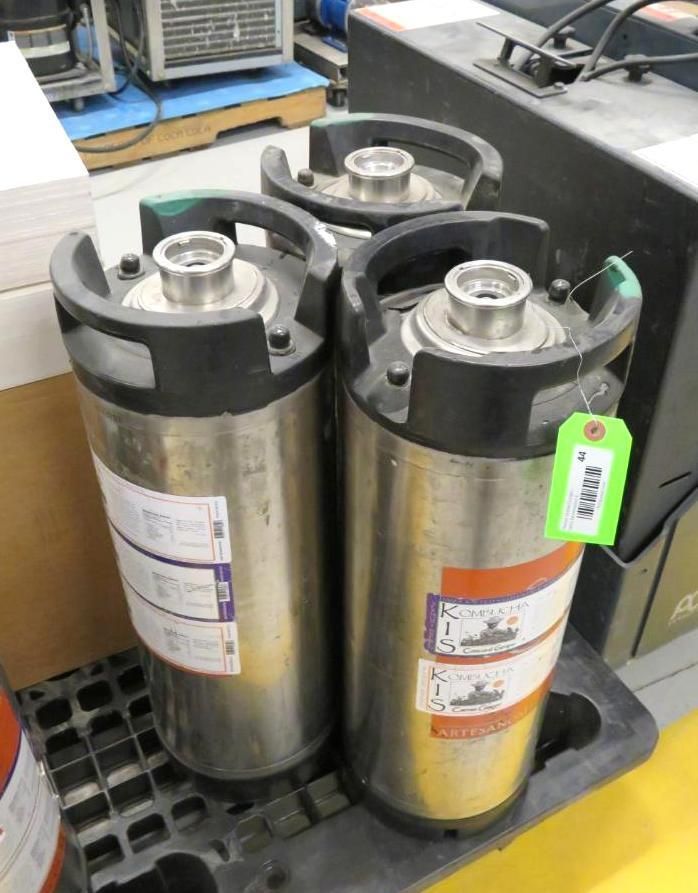 Three stainless steel kegs, possibly for beer, on a black plastic tray. One has a green tag.