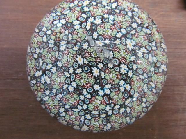 A round glass paperweight with a millefiori pattern of small flowers in white, green, and red on a black background.
