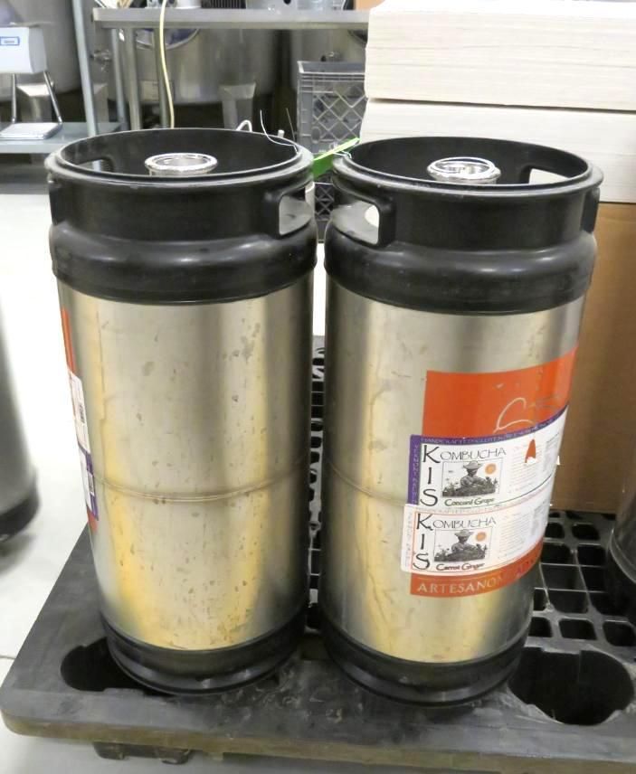 Two stainless steel beer kegs with black tops and labels, resting on a black plastic pallet.