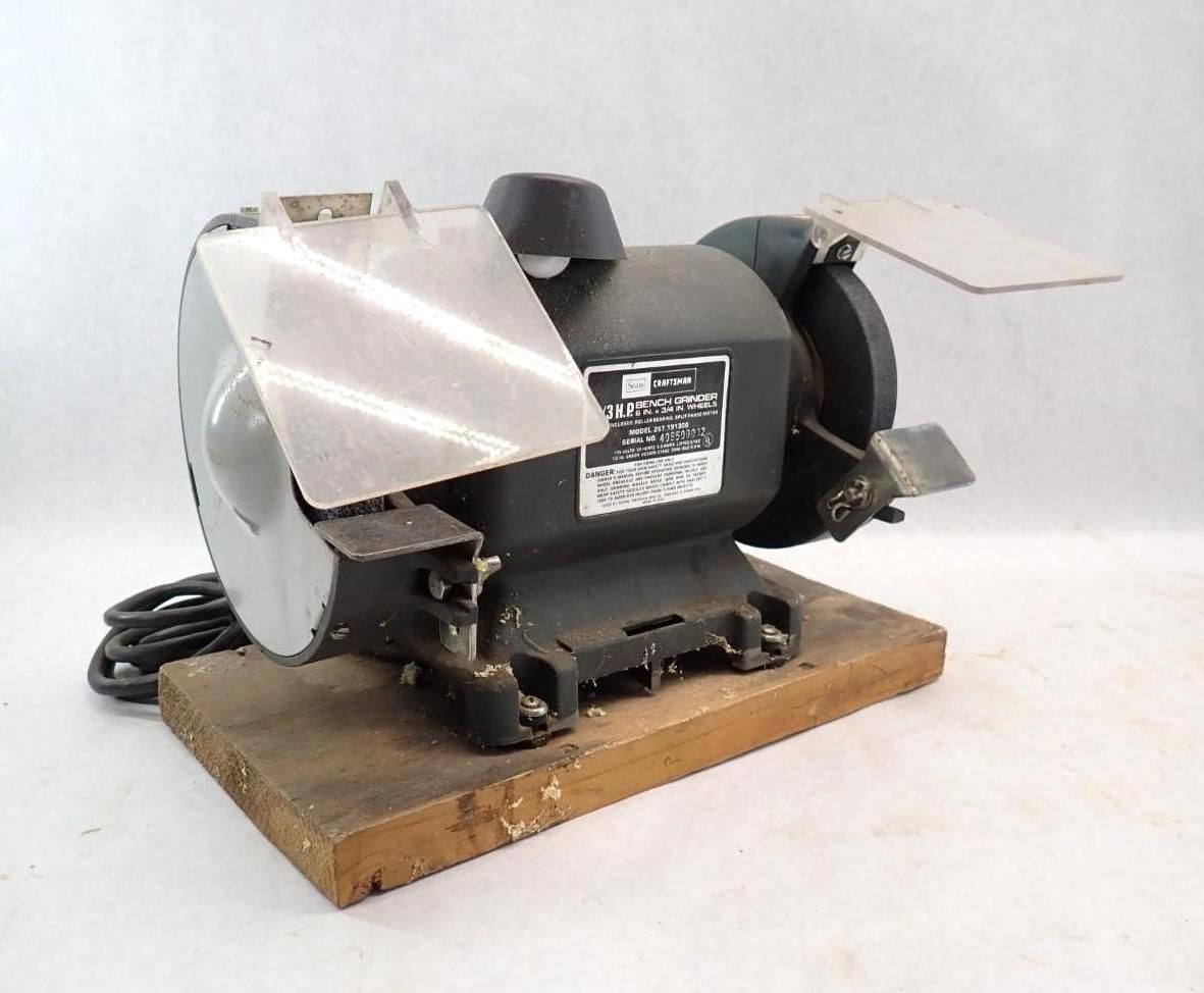 Black bench grinder with protective shields, mounted on a wooden base.