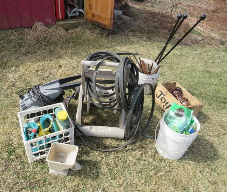 Garden tools and supplies on grass. Includes hose reel, crate, bucket of fertilizer, and wooden box.