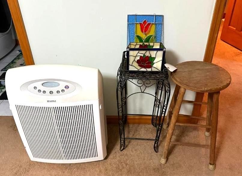 Air purifier next to a decorative stand holding a stained glass box and stool. Beige walls and hardwood floor.
