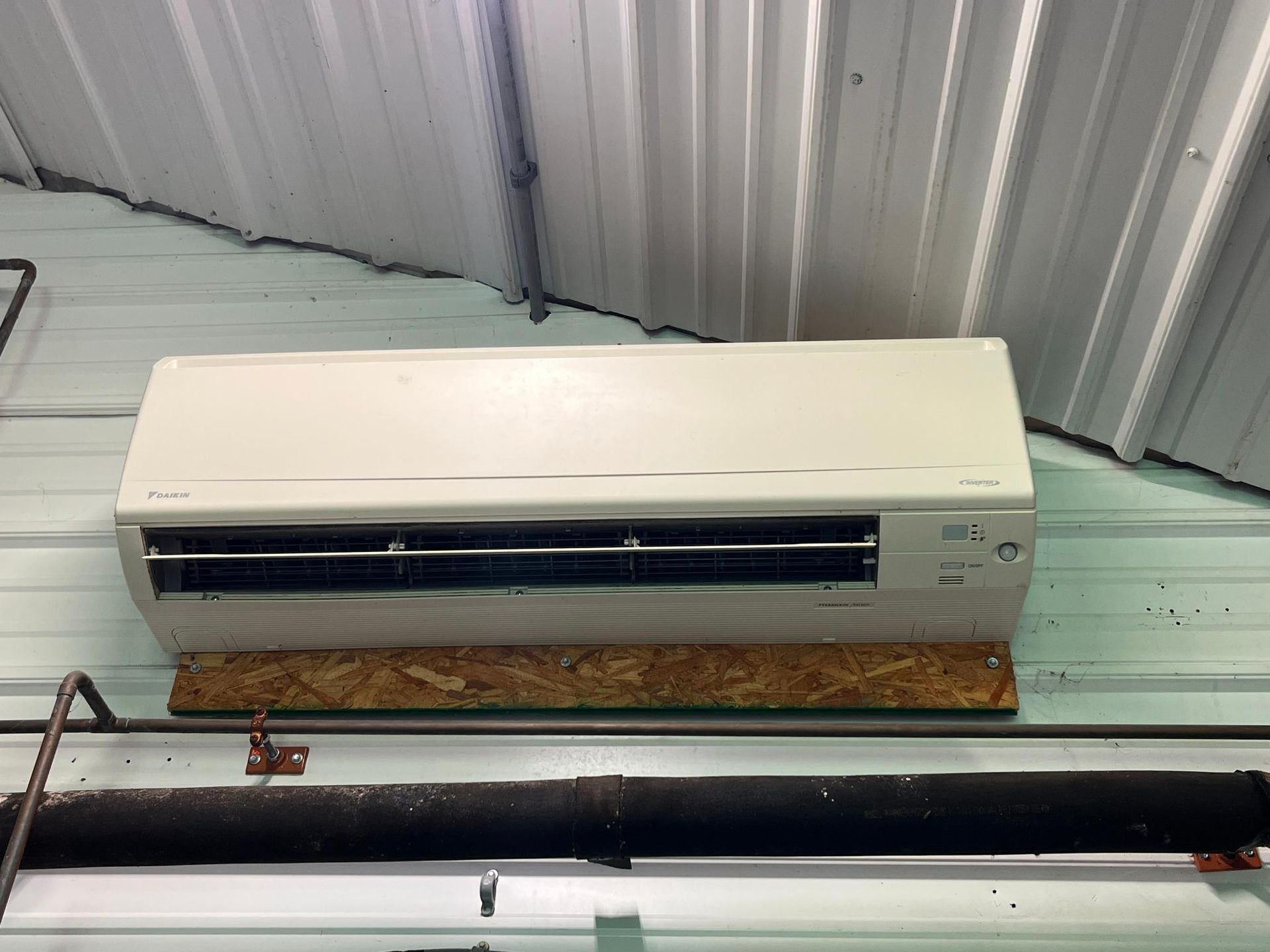 White air conditioner unit mounted on a plywood base, installed on a corrugated metal wall.