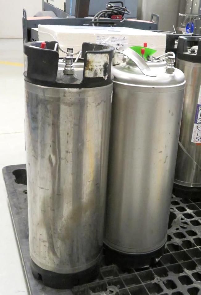 Two metal kegs on a black pallet, one with black top and connectors, the other with a silver lid.
