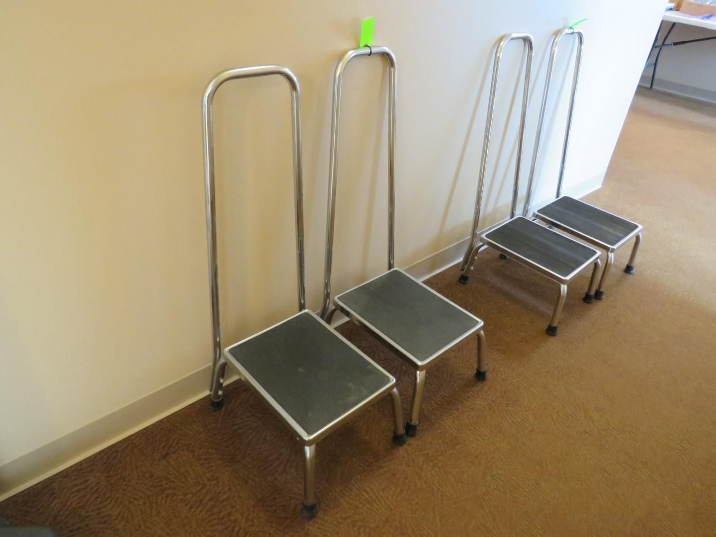 Four metal step stools with handrails against a beige wall and brown carpet.