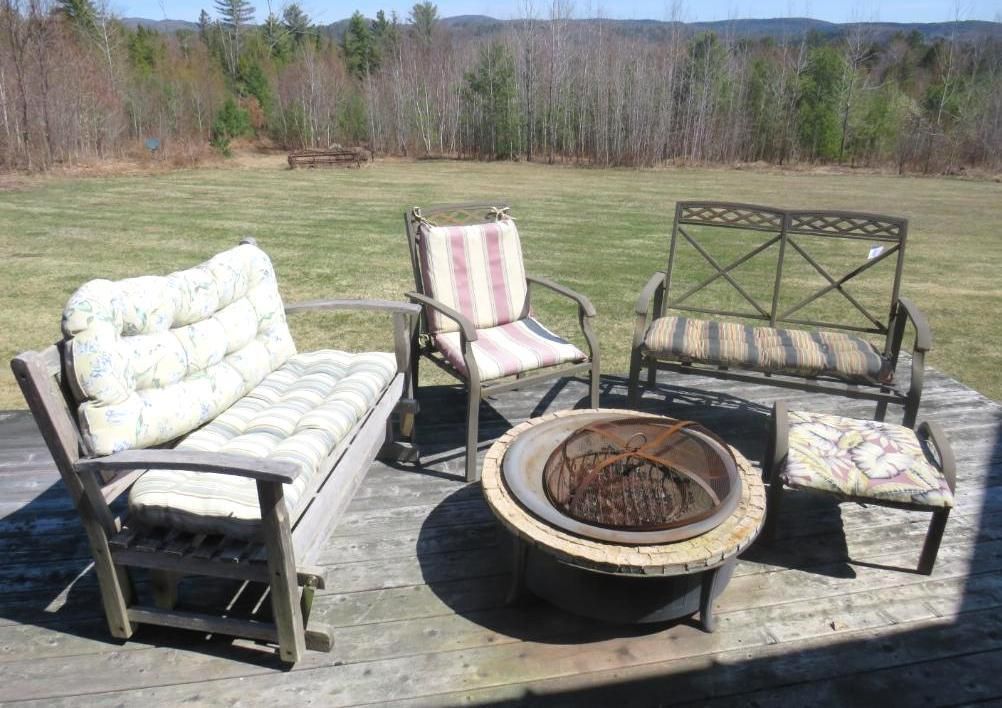 Outdoor seating area with fire pit on wooden deck; chairs, bench, and footstool; grassy field in background.