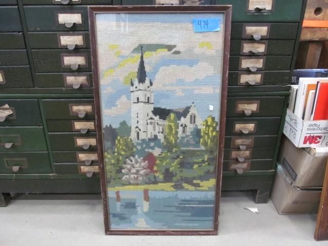Needlepoint of a white church by a lake, framed in brown. Green trees and blue sky.