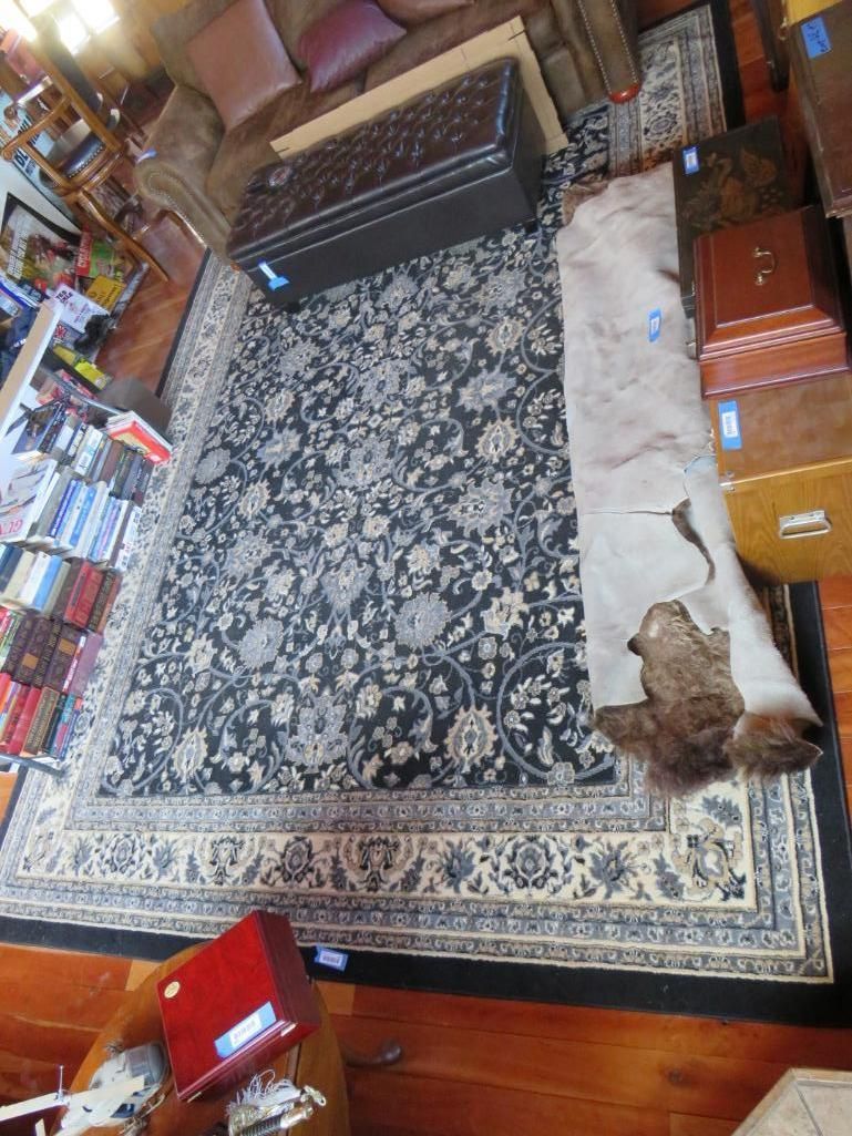 Dark floral rug with ivory border, and a tan hide rug laid on top, in a room with wood floors and furniture.