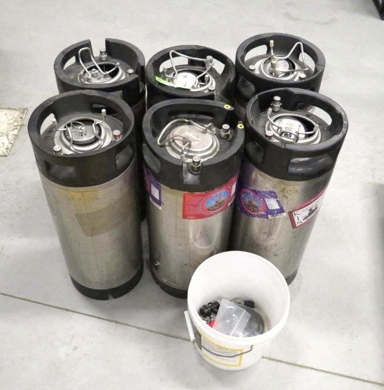 Seven stainless steel kegs with black tops and a bucket of accessories on a concrete floor.