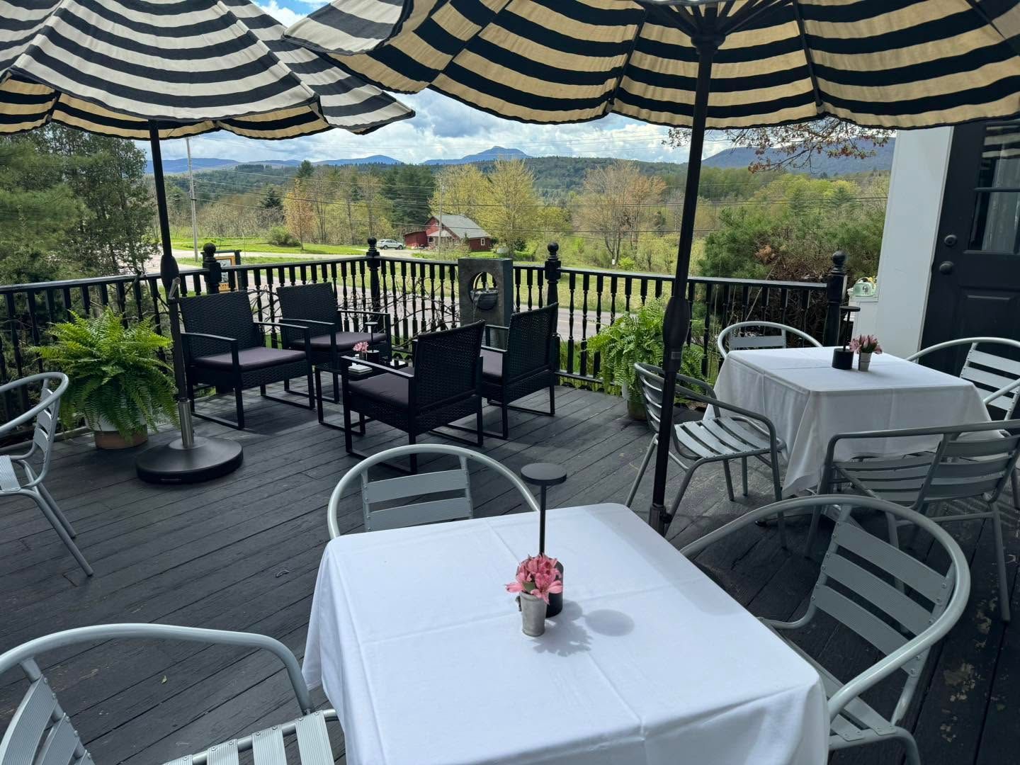 Outdoor café terrace with white tables, black chairs, striped umbrellas, and mountain views
