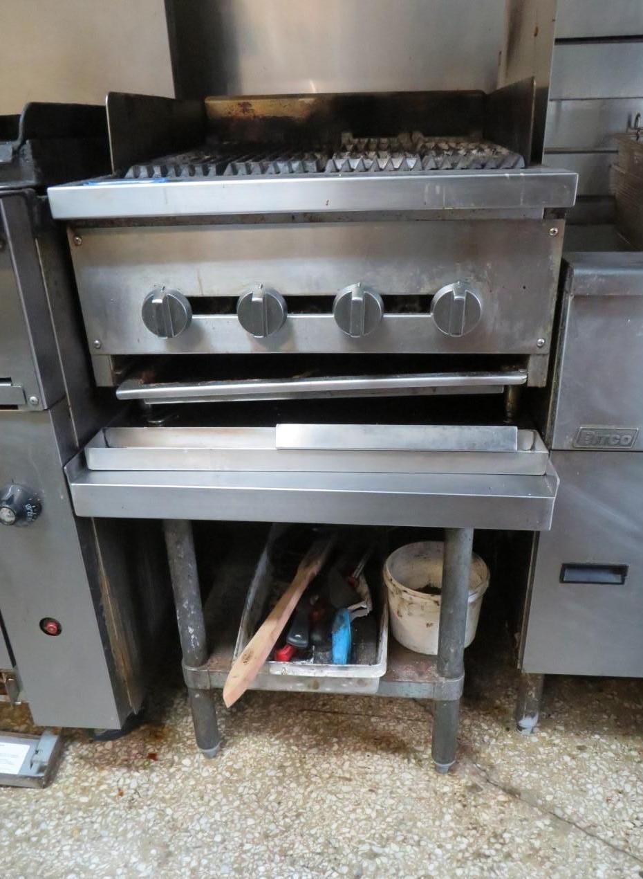 Commercial grill, stainless steel, four knobs, with attached shelving holding tools and a bucket.