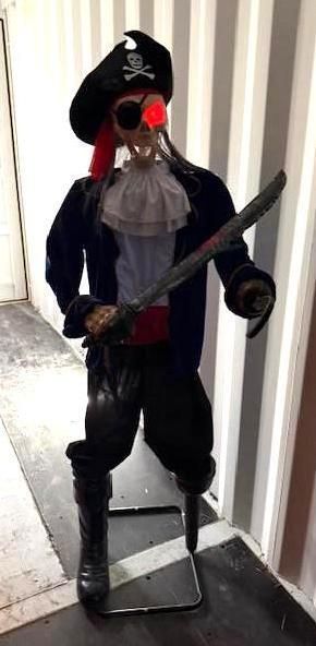 A pirate Halloween decoration with a glowing red eye, sword, and tricorn hat, in a narrow indoor setting.