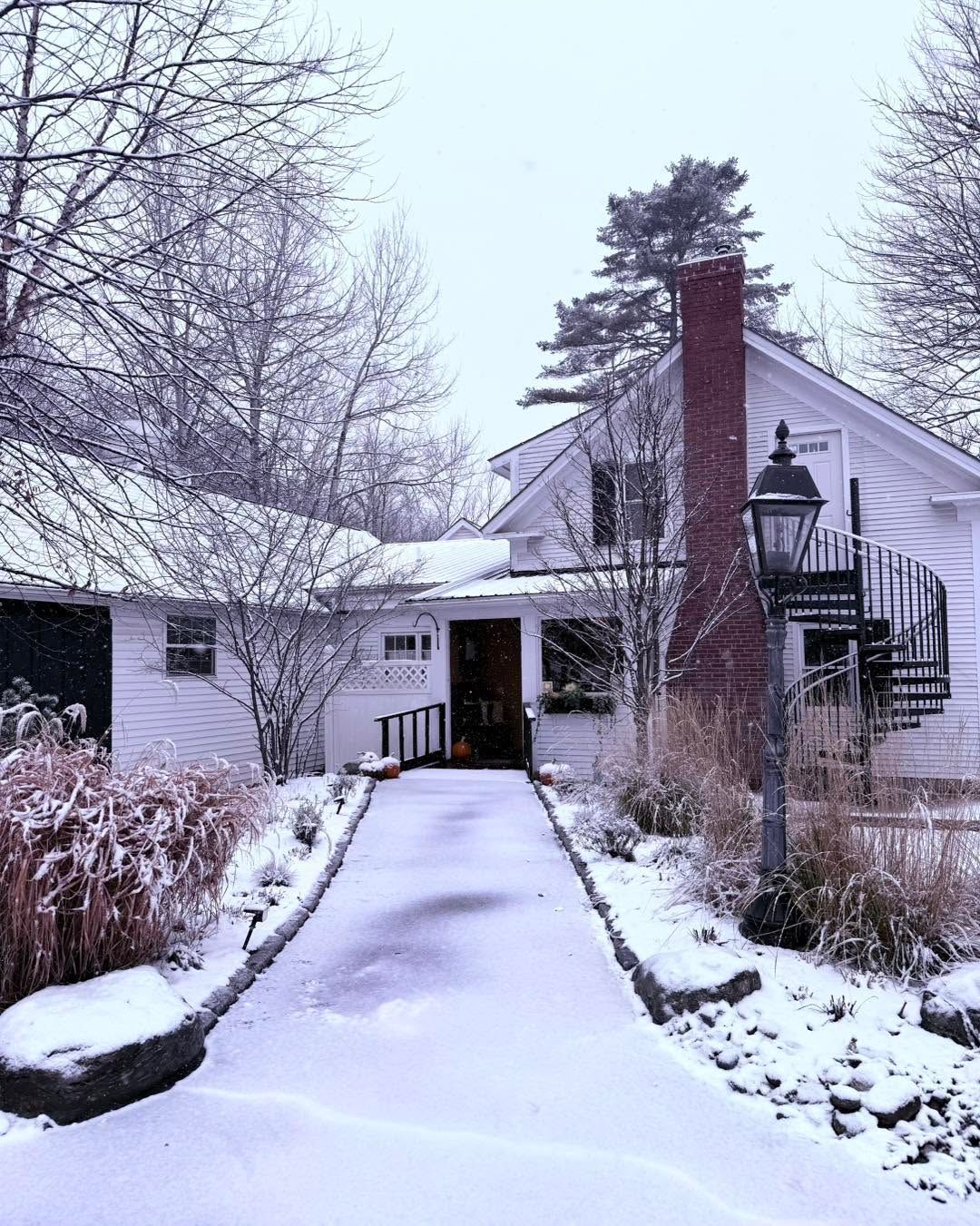 Snow-covered walkway leading to a white house with bare trees and a brick chimney