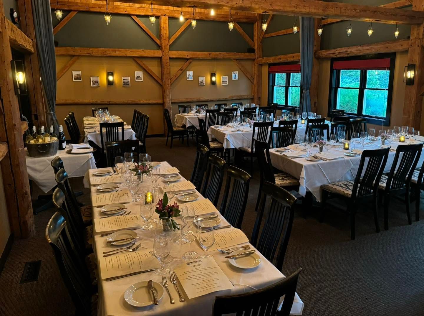 Empty restaurant dining room with set tables, black chairs, warm wood walls, and large windows.