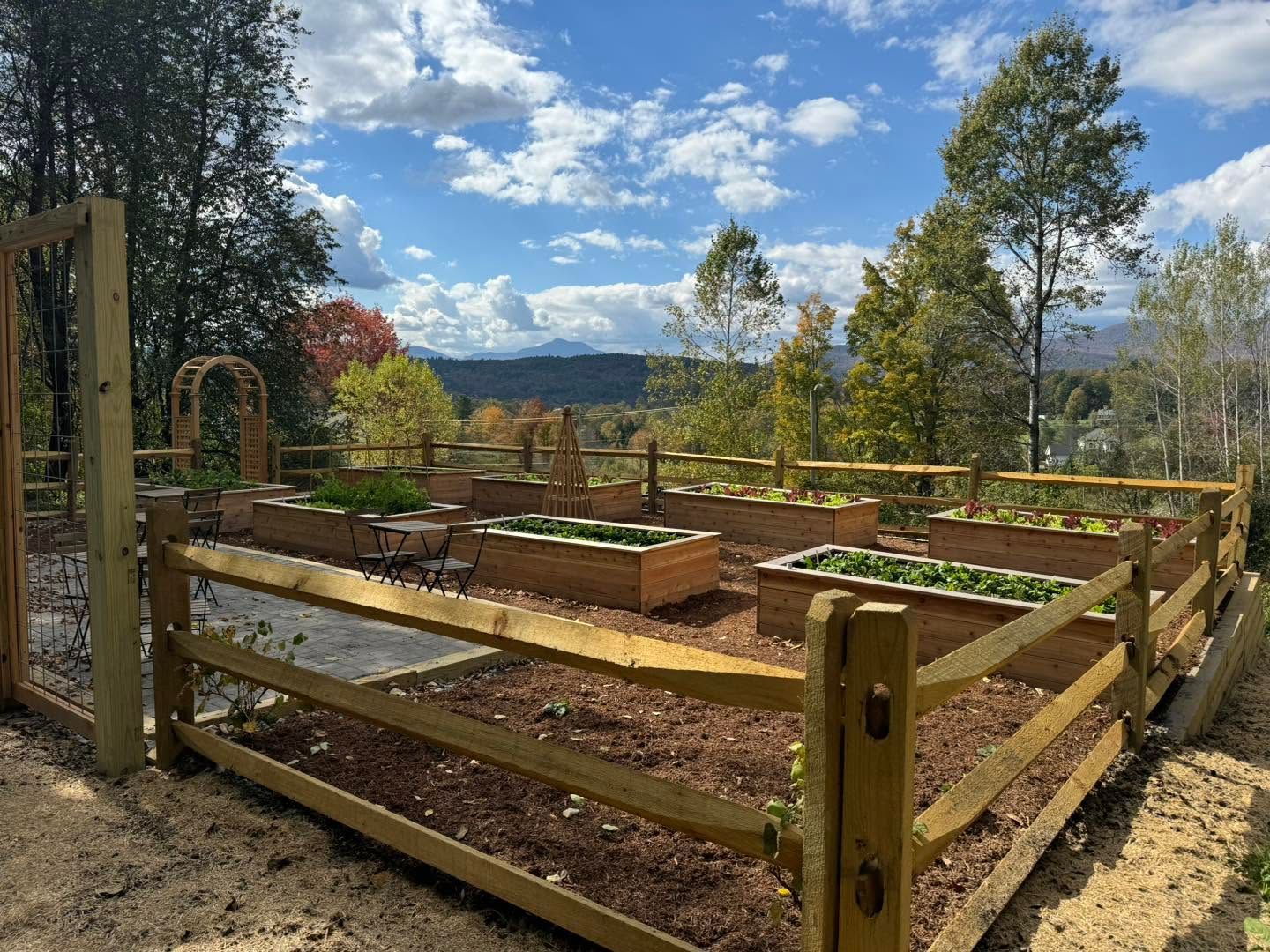 Wooden fenced garden beds on a hilltop with trees and mountains under a partly cloudy sky