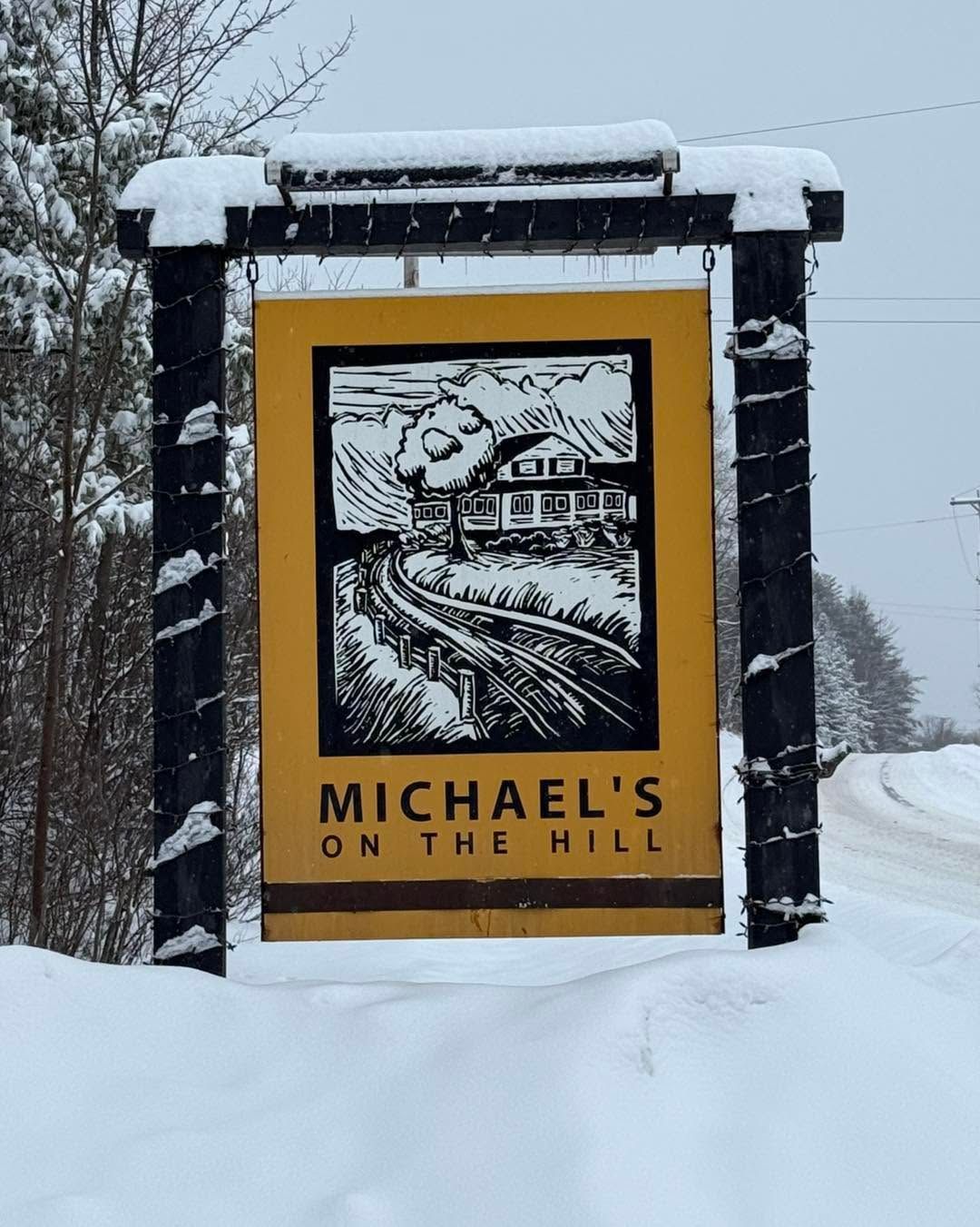 Snowy roadside sign for “Michael’s on the Hill” in yellow and black.