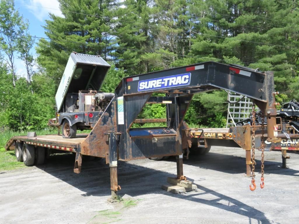 Sure-Trac flatbed trailer in a gravel lot with a raised dump trailer bed behind it, under a bright sky.