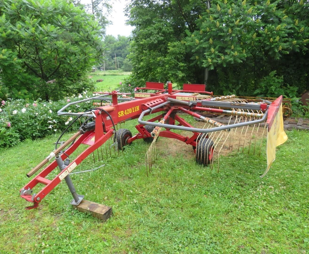 Red farm hay rake on green grass in a field with trees.