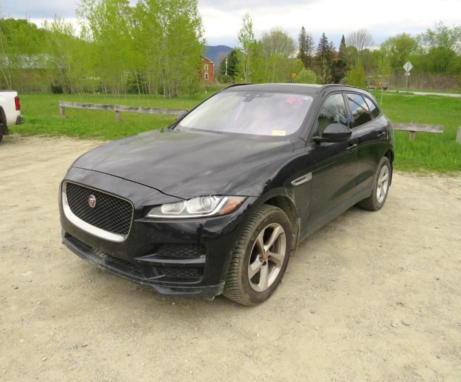 Black Jaguar F-PACE SUV parked outdoors on dirt. Trees and buildings in the background on a cloudy day.