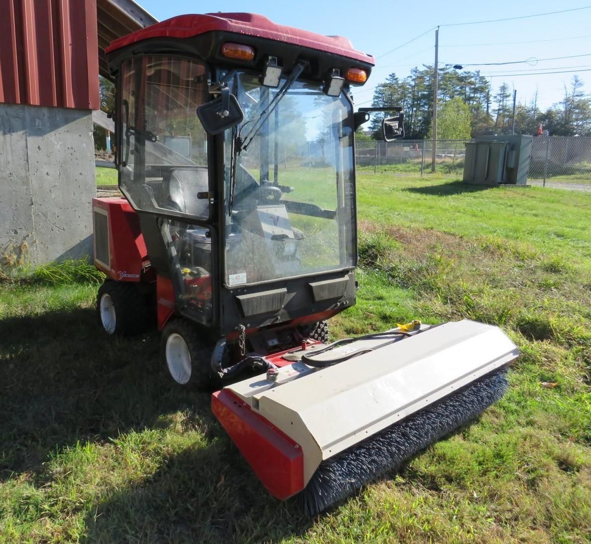 Red and tan compact tractor with enclosed cab and brush attachment, parked on grass.