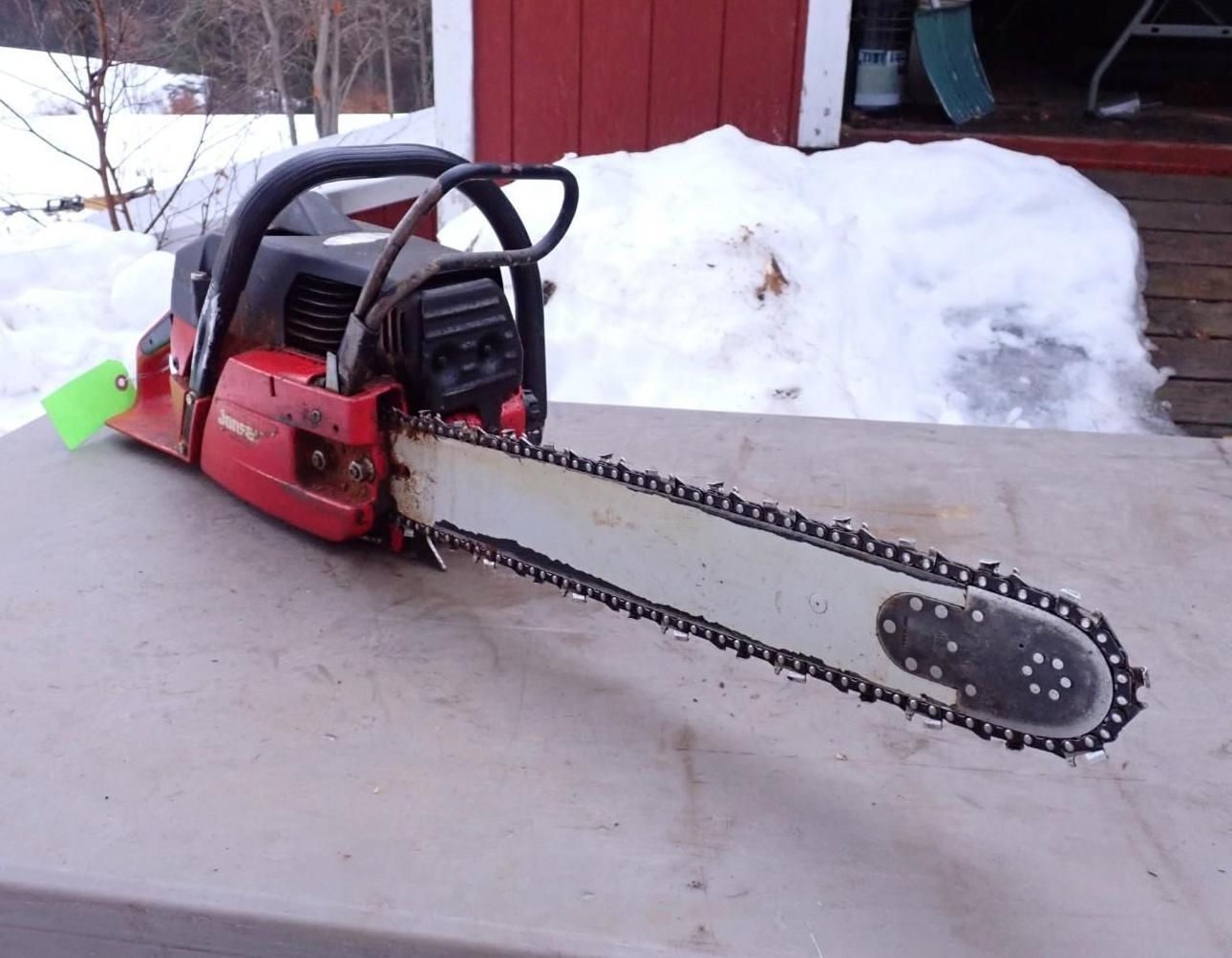 A red gasoline-powered chainsaw rests on a flat surface outdoors against a backdrop of snow and a red building.