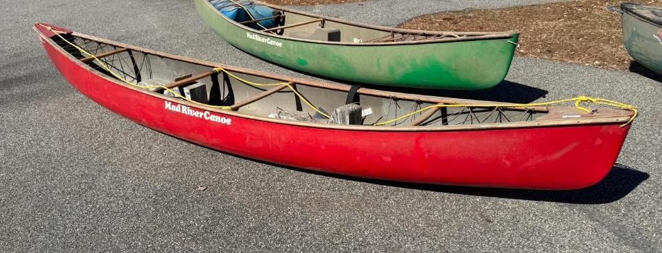 Red canoe in front of a green canoe on a gray surface, outdoors.