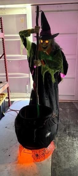 A witch with glowing eyes stirs a cauldron. The scene is lit with orange light, suggesting a Halloween display.