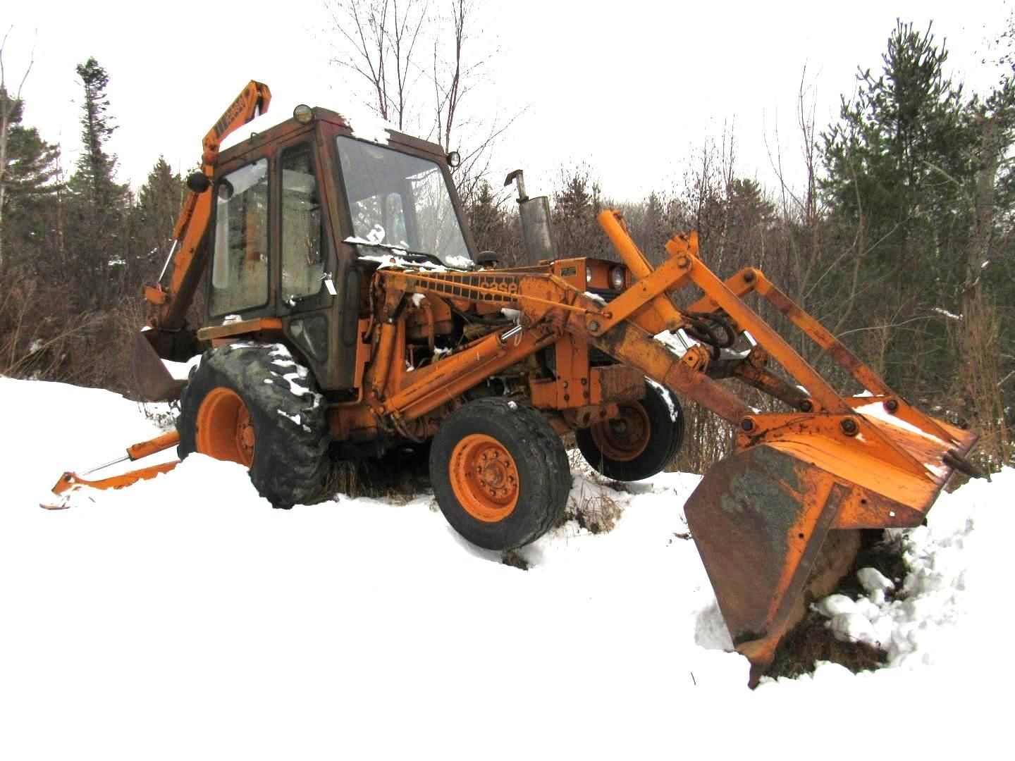 Orange Case 580B Backhoe in snow-covered field. Front bucket and backhoe arm lowered. Overcast day.