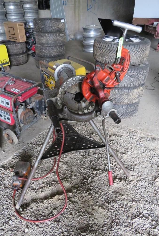 Red pipe threading machine on a metal tripod stand, outdoors. Tires and equipment are in the background.