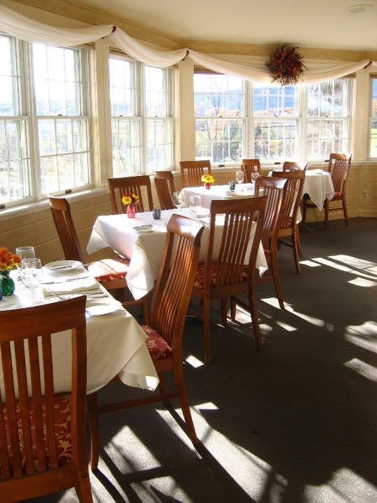 Sunlit dining room with tables set for a meal beside large windows and wooden chairs