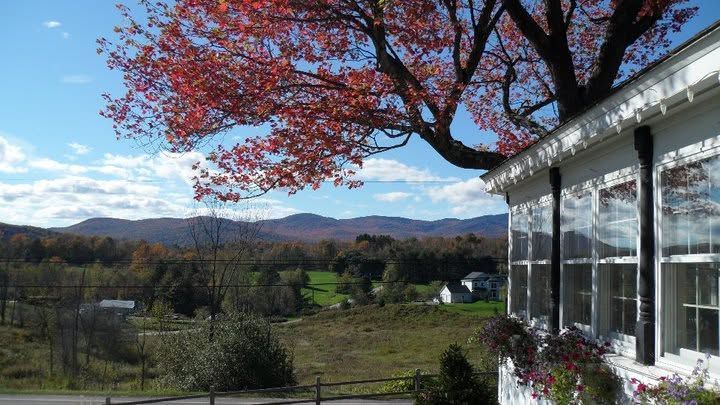 Red maple tree over a rural valley beside a white house under a blue sky