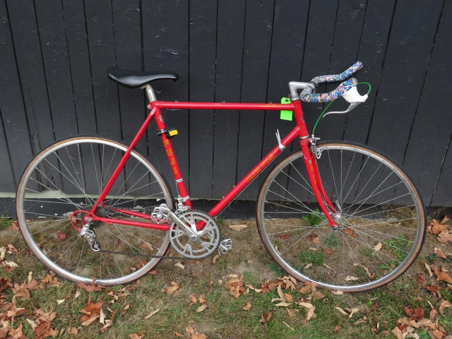 Red road bicycle with drop handlebars and white wheels against a dark wall and grass.