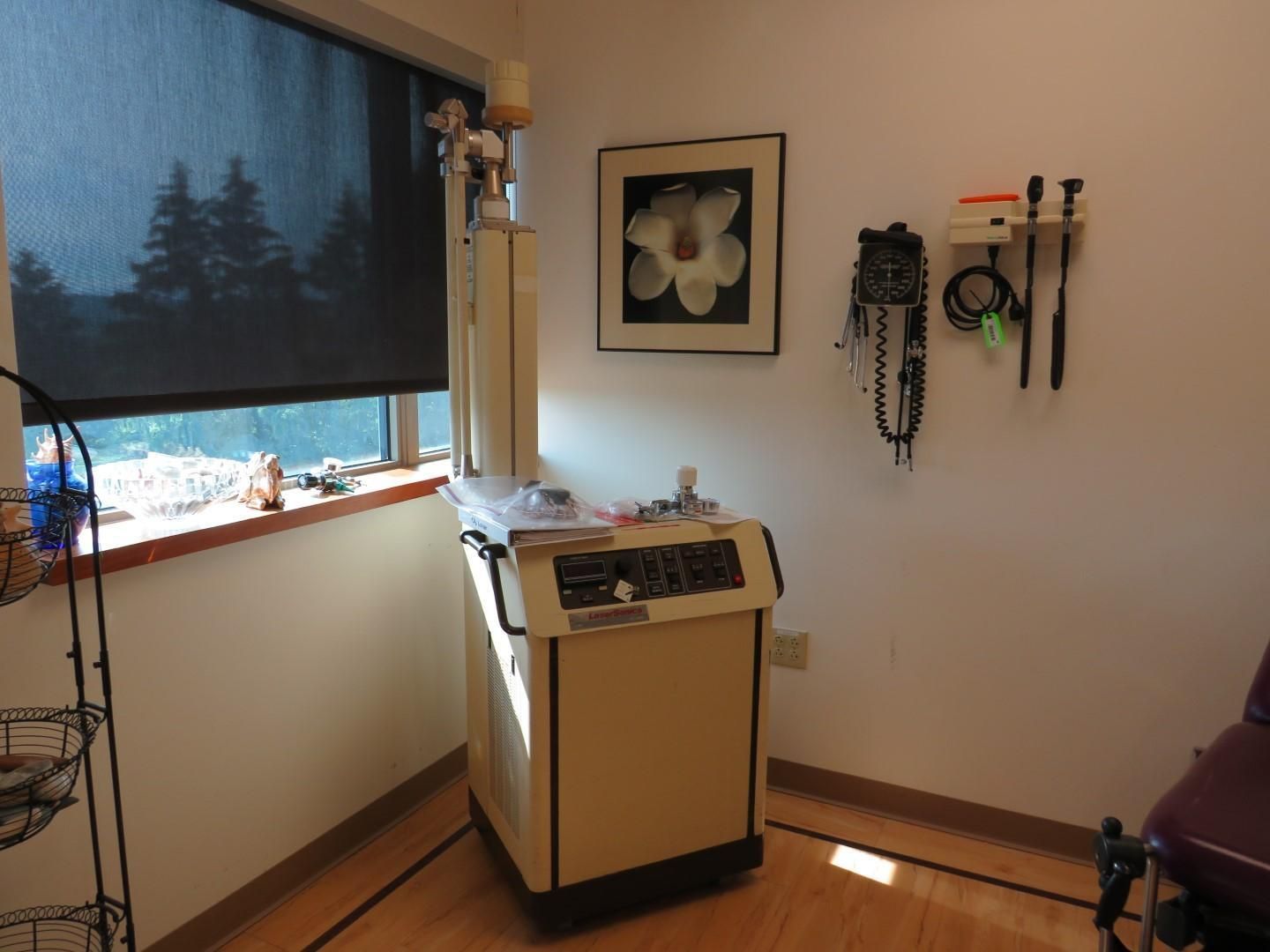 Examination room with medical equipment and window, wood floor, white walls.