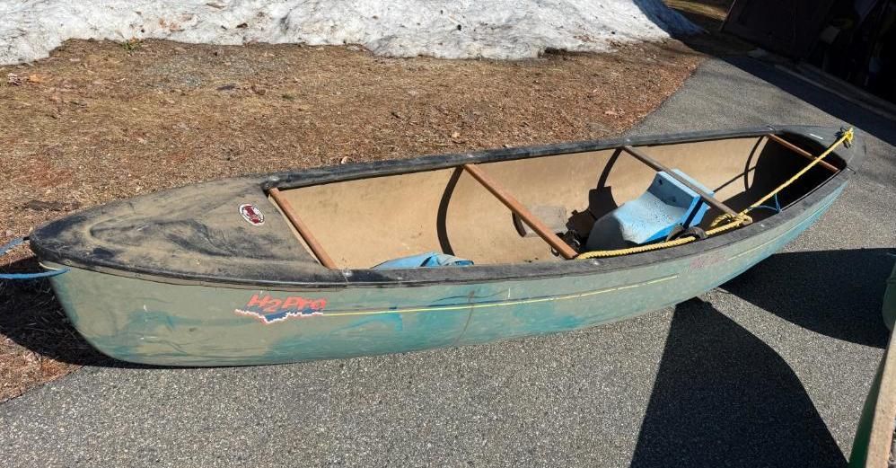 Green canoe on asphalt next to dirt and snow. Interior has seats, rope.