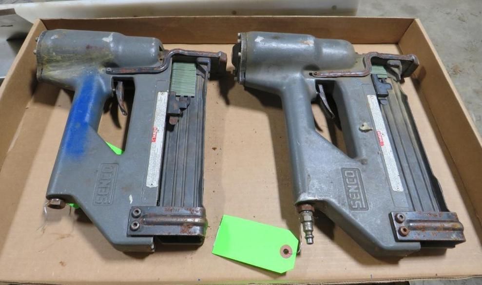 Two grey Senco pneumatic nailers on a cardboard surface. One has a blue grip, the other a green tag.