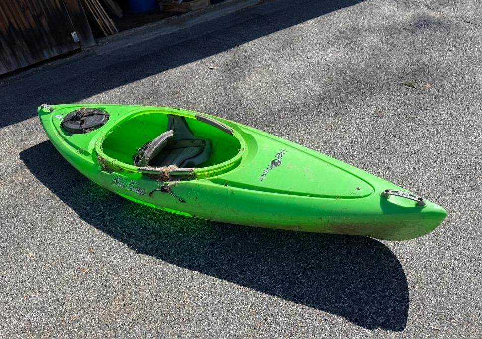 Green kayak on an asphalt surface, with an open cockpit and a dark circular hatch.