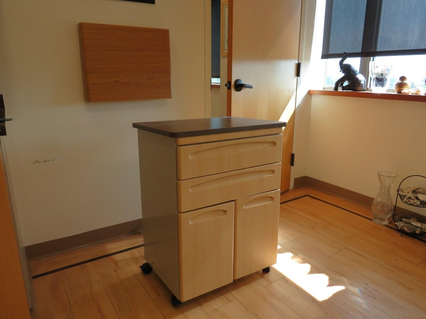 A light wood cabinet on wheels, with drawers and doors, in a room with a window and a door.