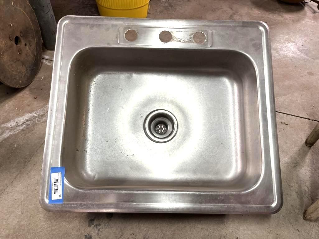 Stainless steel kitchen sink with faucet holes, resting on a concrete floor.