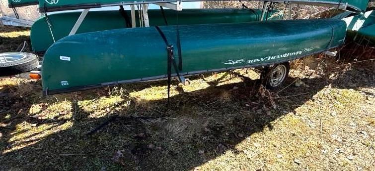 Green canoe on a small trailer, secured with straps, parked on dry grass.